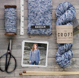 blue white aran weight skein machine washable for knitting crocheting and weaving some wound up of a spindle a measuring tape a knitted swatch a skein a pair of scissors a photo of a white woman with ginger hair wearing a jumper made of the yan a pair of knitting needles  