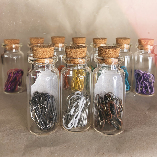 ten clear glass bottles with cork tops seven in the back three in the front silver white grey brown steel pink purple blue green yellow orange stitch markers 