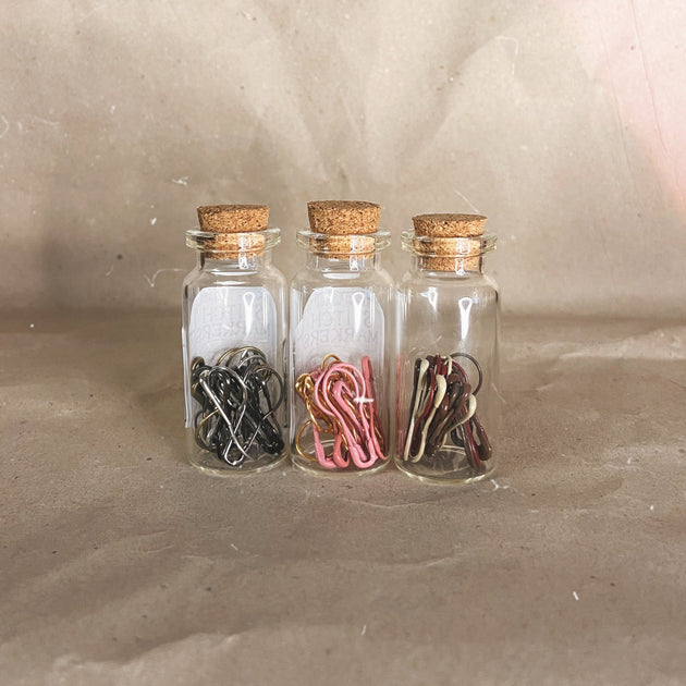 three small glass bottles with a cork top silver black gold pink white red brown stitch markers in 