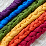 purple blue green yellow orange red eco nylon biodegradable braids lined up in a row on a white crocheted background 