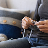 white woman sitting knitting with chunky weight grey yarn knitted pillow in the background