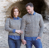 a white woman with ginger hair and a white man with light brown hair both wearing matching knitted jumpers made of orange blue green white aran weight yarn