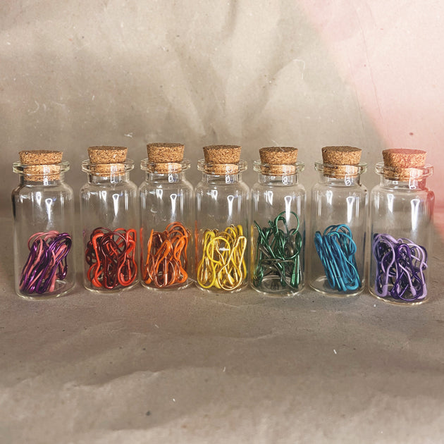 seven small clear bottles with a cork top pink red orange yellow green blue purple stitch markers 